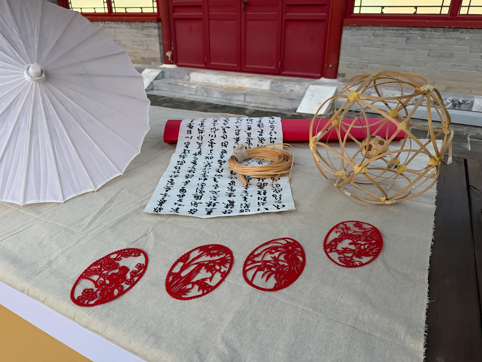 Red paper cuttings, a bamboo lantern, calligraphy and a white umbrella arranged on a linen-covered table.