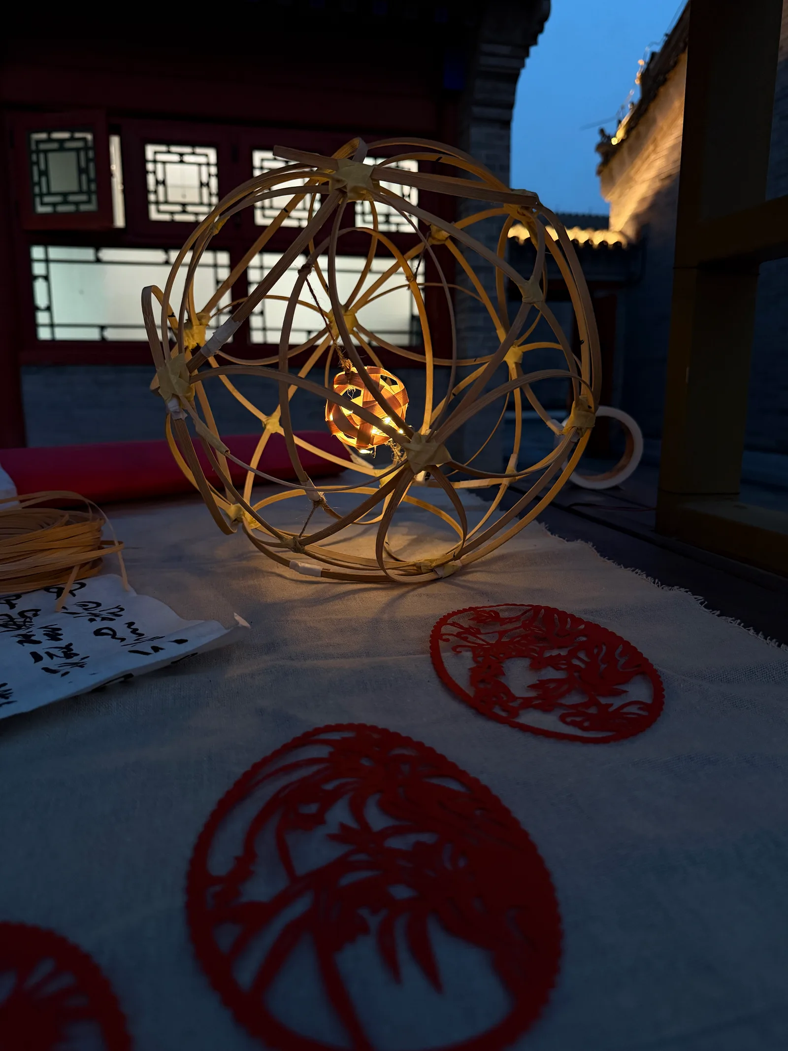 A bamboo lantern glowing beside red paper cuttings at dusk.